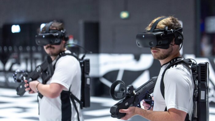 Two men in white shirts hold VR guns and wear VR headsets during a zombie shooting game