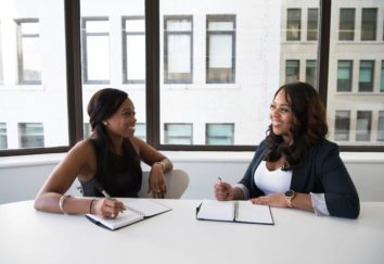 Two women sit in an office in smart clothing smiling at one another