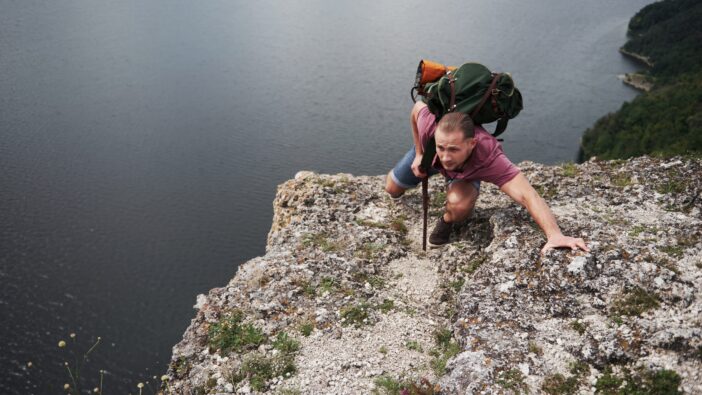 A man climbs a tall rock above a vast body of water wearing a backpack filled with supplies