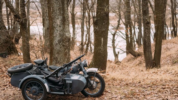An old motorcycle rests quietly among the trees in a serene woodland setting, showcasing its vintage charm.